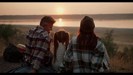 Rear-view-of-a-happy-married-couple-along-with-their-little-daughter-sitting-on-a-mat-during-their-picnic-and-enjoying-the-sunset-view-near-a-pond-in-the-summer-outside-the-city-in-the-evening