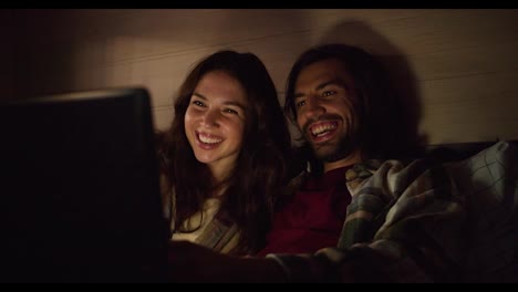 A-happy-couple,-a-brunette-guy-with-stubble-and-his-brunette-girlfriend-in-a-plaid-shirt,-lie-on-the-sofa-in-a-trailer-and-watch-a-funny-comedy-using-a-laptop-with-dimmed-lights-in-an-atmospheric-romantic-evening-near-a-camp-in-a-trailer-outside-the-city-in-the-summer