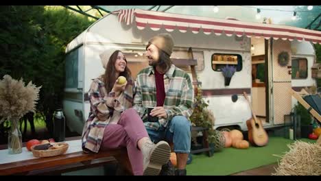 A-happy-brunette-girl-in-a-white-hat-holds-a-green-apple-in-her-hand-and-communicates-with-her-brunette-boyfriend-in-a-green-checkered-shirt-who-drinks-tea-and-sits-near-a-trailer-in-a-camp-outside-the-city-in-the-summer-during-a-picnic