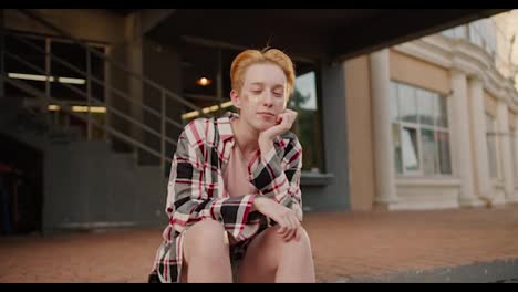 Portrait-of-a-lesbian-girl-with-a-short-haircut-with-sparkles-on-her-face-in-a-pink-checkered-shirt-who-sits-on-a-step-on-the-street,-propping-her-head-up-with-her-palm,-looking-at-the-camera