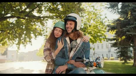 Happy-couple-brunette-girl-in-a-plaid-shirt-leans-and-hugs-her-boyfriend-with-curly-hair-in-a-White-moped-helmet-and-denim-jacket,-who-sits-on-a-moped-in-the-summer-city