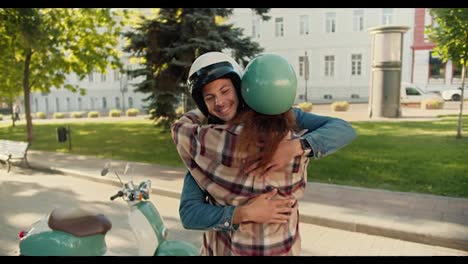 Happy-guy-with-long-curly-hair-in-a-denim-shirt-and-a-white-motorcycle-helmet-hugs-his-brunette-girlfriend-in-a-checkered-shirt-Near-his-motorcycle-in-a-summer-city-park