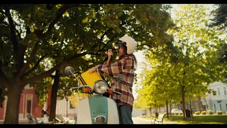 A-brunette-courier-girl-in-a-checkered-shirt-wearing-a-white-motorcycle-helmet-and-with-a-yellow-bag-comes-up-and-puts-it-on-her-moped.-A-girl-inspects-an-order-that-she-is-going-to-deliver-on-the-street-in-the-summer