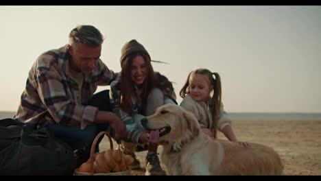 Close-up-shot-of-a-happy-cream-colored-dog-resting-on-a-deserted-seashore-with-its-owners.-A-happy-brunette-man-and-his-wife-in-a-green-checkered-shirt-are-stroking-their-dog-and-their-little-daughter-is-sitting-near-them-during-a-picnic-and-hike-in-the-summer