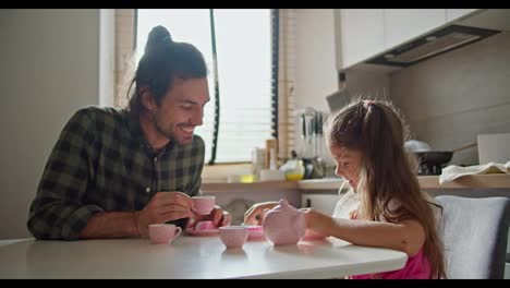 Happy-brunette-man-in-a-green-checkered-shirt-together-with-his-little-daughter-brunette-girl-in-a-pink-dress-play-tea-party-using-toy-pink-cups-and-a-teapot-in-the-kitchen