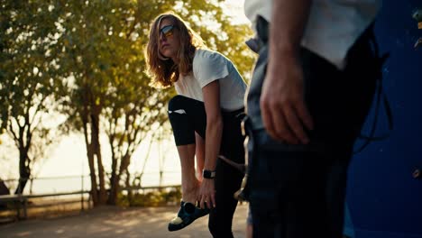 A-cool-blonde-girl-climber-in-a-white-T-shirt-and-black-sunglasses-puts-on-a-special-uniform-and-equipment-with-her-friend-before-starting-a-rock-climbing-lesson-at-a-climbing-wall