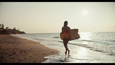 Back-view:-A-blonde-girl-in-an-orange-swimsuit-carries-a-light-colored-wooden-surfboard-and-walks-along-the-sandy-seashore-in-the-morning-at-Sunrise.-A-blonde-girl-surfing-master-walks-along-the-sea-with-her-surfboard