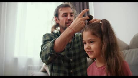 A-brunette-guy-with-long-hair-in-a-green-checkered-shirt-gives-his-little-daughter-in-a-pink-jacket-a-ponytail-hairstyle-using.-Brunette-man-Single-father-doing-his-daughter's-hair-using-a-special-elastic-band-in-a-modern-home-Sitting-on-the-sofa