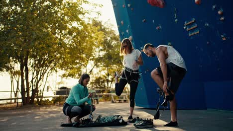 Two-girls-in-sports-uniforms-and-a-guy-in-a-white-T-shirt-put-on-special-equipment-and-insurance-before-climbing-at-a-climbing-wall.-A-group-of-climbers-warm-up-before-climbing-the-climbing-wall-on-a-sunny-summer-day