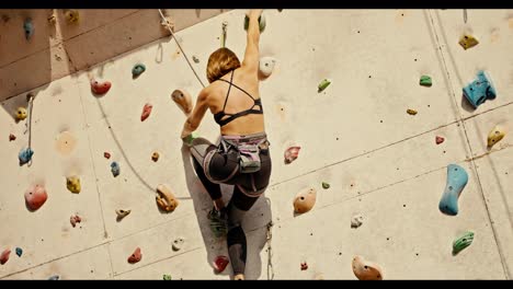 A-blonde-girl-with-a-bob-hairstyle-in-a-black-top-and-black-pants-with-the-necessary-equipment-and-insurance-climbs-up-the-route-of-a-climbing-wall-on-a-sunny-summer-day