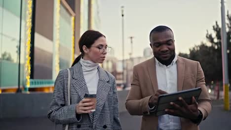 A-confident-male-manager-with-Black-skin,-a-short-haircut-and-a-beard-in-a-brown-suit-walks-and-talks-with-his-colleague-a-businesswoman-a-brunette-in-round-glasses-and-a-gray-coat,-and-looks-at-something-on-a-Black-tablet-during-his-business-meeting-in-the-city