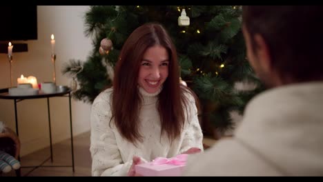 Over-the-shoulder,-a-guy-gives-his-brunette-girlfriend-in-a-white-Christmas-sweater-a-gift-in-a-pink-box-with-a-bow-and-the-girl-opens-this-gift-near-the-Christmas-tree-in-the-evening-in-a-cozy-home