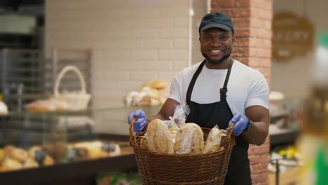 Portrait-of-a-happy-man-in-a-black-cap-and-white-T-shirt-in-a-black-apron-who-holds-in-his-hands-a-large-basket-of-pastries-in-a-modern-grocery-store