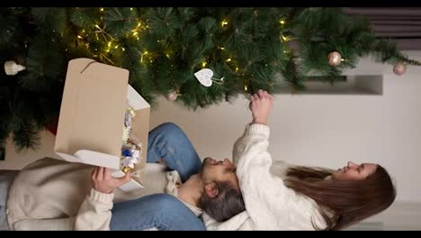 Vertical-video:-of-a-guy-holding-a-box-with-Christmas-decorations-in-his-hands-and-a-girl-sitting-on-his-shoulders-taking-decorations-from-the-box-and-attaching-them-to-a-Green-Christmas-tree