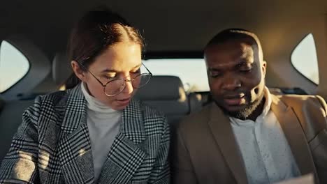 A-confident-brunette-girl.-A-businessman-with-round-glasses-in-a-gray-jacket-communicates-with-his-colleague-a-man-with-Black-skin-and-a-beard-in-a-brown-jacket-during-his-business-trip-in-the-cabin-of-a-modern-car-outside-the-city