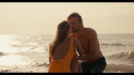 A-blonde-girl-in-an-orange-swimsuit-kisses-her-blonde-boyfriend-with-a-bare-torso-in-black-shorts-while-sitting-on-the-beach-near-the-sea-early-in-the-morning