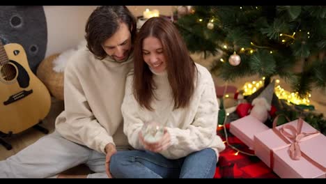 Happy-couple,-a-brunette-guy-in-a-white-sweatshirt-and-a-brunette-girl-in-a-White-sweater-who-shakes-the-New-Year's-Sphere-and-looks-at-the-flying-snow-near-pink-gifts-of-a-red-blanket-and-a-green-Christmas-tree