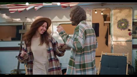 Happy-brunette-girl-in-light-brown-together-with-her-brunette-boyfriend-in-a-green-checkered-shirt-near-their-trailer-in-the-camp-during-a-picnic-outside-the-city-in-summer