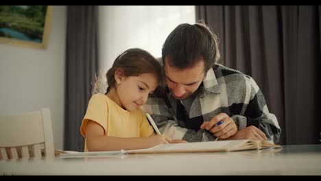 A-little-brunette-girl-in-a-yellow-dress-does-her-homework-and-writes-in-her-notebook-together-with-her-dad,-a-brunette-man-in-a-checkered-shirt-on-a-white-table-in-a-room-in-a-modern-apartment