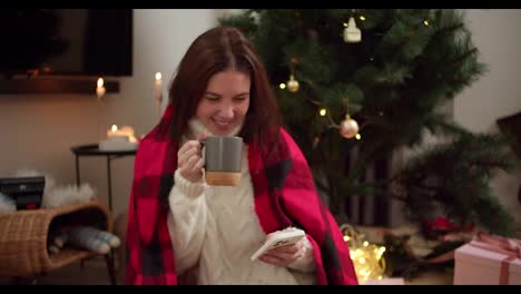 Happy-brunette-girl-in-a-red-plaid-and-white-sweater-drinks-hot-tea-from-a-gray-mug-and-works-on-her-white-phone-near-the-Christmas-tree-in-a-cozy-decorated-room