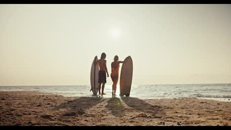 Rear-view-from-afar-of-a-brunette-guy-together-with-his-blonde-girlfriend-in-an-orange-swimsuit-standing-and-looking-towards-the-sea-and-holding-their-surfboards-near-them-on-the-orange-sandy-beach-in-the-morning