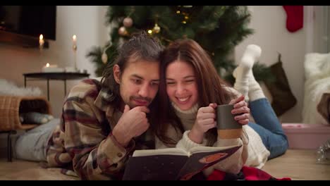 Interested-guy-and-girl-lying-on-the-floor-and-reading-a-Christmas-book-along-with-a-cup-of-hot-drink-in-a-cozy-room-with-a-Christmas-tree-and-a-decorated-room-in-winter-evening