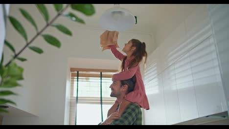 A-little-brunette-girl-in-a-pink-dress-sits-on-the-shoulders-of-her-brunette-dad-in-a-green-checkered-shirt-and-wipes-the-lamp-on-the-ceiling-with-a-special-cloth-in-a-modern-kitchen