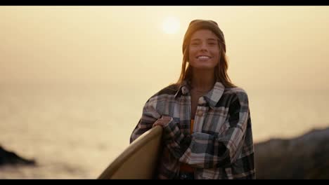 Portrait-of-a-blonde-girl-in-a-black-hat-and-plaid-shirt-who-holds-a-surfboard-stands-on-a-rocky-shore-near-the-sea-in-the-morning-and-looks-at-the-camera