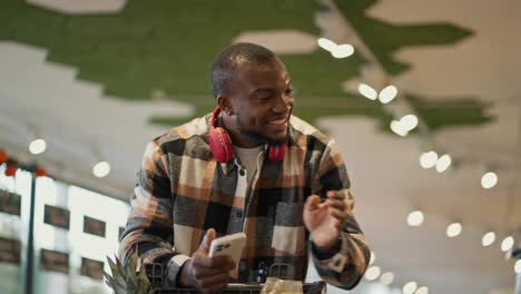 A-happy-man-with-Black-skin-in-a-checkered-shirt-and-red-wireless-headphones-leaning-on-a-cart-drives-through-the-supermarket-and-examines-the-goods-on-the-counter-and-selects-from-them-those-that-he-needs