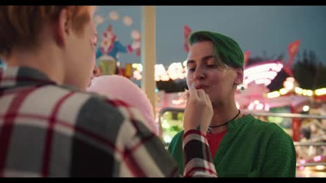 A-blonde-girl-with-a-short-haircut-in-a-plaid-shirt-feeds-her-girlfriend-with-a-short-green-haircut-in-a-Green-shirt-and-also-passes-a-piece-of-pink-cotton-candy-with-her-mouth-during-their-date-in-a-bright-and-glowing-amusement-park