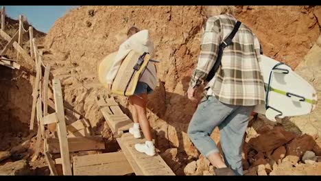 A-blond-guy-in-a-plaid-shirt-and-his-blonde-girlfriend-in-a-white-sweatshirt-climb-a-wooden-ladder-on-a-rocky-beach-and-carry-surfboards-with-them