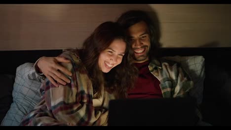 Happy-couple-watching-a-movie-with-dim-lights-in-a-trailer-outside-the-city-in-summer.-Happy-brunette-girl-in-a-pink-checkered-shirt-lies-with-her-brunette-boyfriend-in-a-green-checkered-shirt,-who-hugs-her-while-watching-a-movie-together-on-a-laptop-in-a-camp-in-a-trailer-in-the-summer-evening