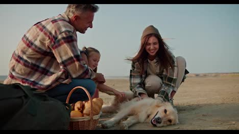 A-happy-brunette-girl-in-a-hat-in-a-green-checkered-shirt,-together-with-her-husband,-strokes-a-large-cream-colored-dog-and-near-them-their-little-daughter-sits-and-relaxes-during-their-picnic-on-the-deserted-seashore-in-the-summer