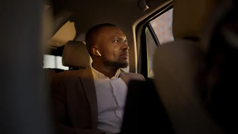 A-confident-man-with-Black-skin-in-a-brown-suit-listens-to-music-on-wireless-headphones-and-looks-out-the-window-at-a-sunny-evening-during-his-business-trip-in-the-interior-of-a-modern-car