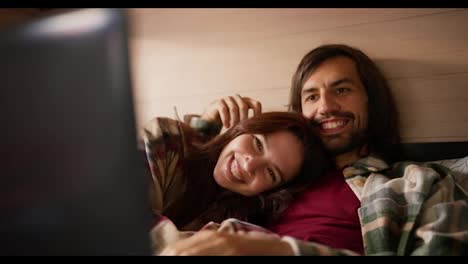 A-happy-brunette-girl-in-a-checkered-shirt-lies-on-the-chest-of-her-brunette-boyfriend-with-stubble-in-a-Green-checkered-shirt-and-they-watch-a-movie-together-using-a-laptop-lying-on-the-sofa-in-a-trailer-in-the-summer-outside-the-city-in-the-camp