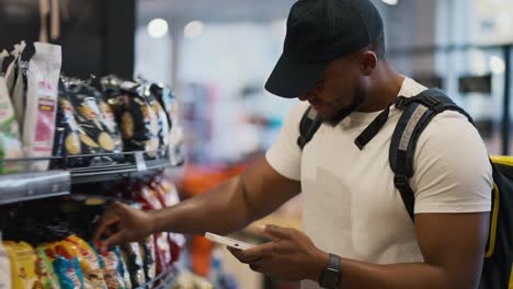 Un-Hombre-Feliz-Y-Confiado,-De-Piel-Negra,-Con-Una-Camiseta-Blanca-Y-Una-Gorra-Negra,-Un-Repartidor-De-Comida-Elige-Los-Productos-Necesarios-Y-Revisa-La-Lista-Mientras-Sostiene-Un-Teléfono-En-Sus-Manos-Y-Una-Gran-Bolsa-Amarilla-Sobre-Sus-Hombros.