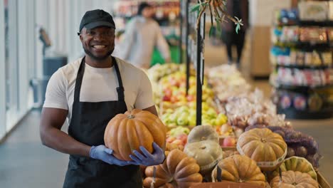 Portrait-of-a-happy-man-with-Black-skin-color-in-a-white-T-shirt-near-a-display-case-with-pumpkins-a-man-holds-a-pumpkin-in-his-hands-poses-and-smiles-broadly-in-a-large-grocery-store