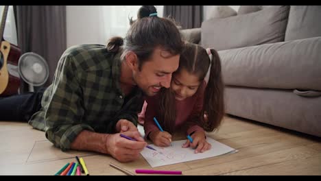 A-lonely-father,-a-brunette-man-in-a-green-checkered-shirt-lies-on-the-floor-with-his-little-daughter-in-a-pink-dress-and-draws-on-a-sheet-of-paper-using-multi-colored-felt-tip-pens-near-a-gray-sofa-in-a-modern-apartment