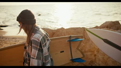 Close-up-shot-of-a-brunette-guy-in-a-white-T-shirt-and-a-blonde-girl-in-a-checkered-shirt-walking-along-the-rocky-seashore-and-carrying-surfboards-in-the-morning-at-sunrise