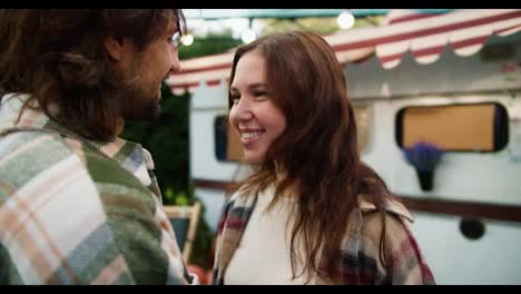 Over-the-shoulder,-a-happy-brunette-girl-in-a-pink-checkered-shirt-hugs-her-brunette-boyfriend-in-a-green-checkered-shirt-and-kisses-her-on-the-forehead-during-her-vacation-and-picnic-outside-the-city-in-a-camp-in-the-summer