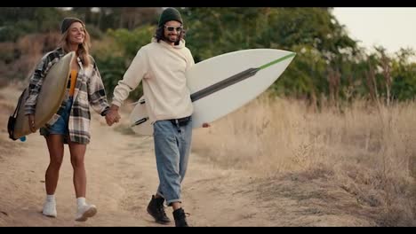 A-happy-guy-in-sunglasses-in-a-white-sweatshirt-walks-with-his-blonde-girlfriend-in-a-plaid-shirt-with-surfboards-in-his-hands-along-a-path-along-dry-summer-grass-and-small-trees