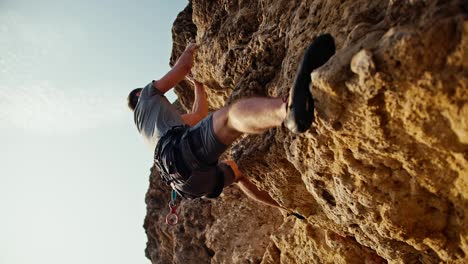 Shot-from-below,-a-man-in-a-gray-T-shirt-and-gray-pants-in-a-special-insurance-climbs-a-yellow-steep-rock-and-stones-against-the-backdrop-of-a-sunny-clear-summer-sky