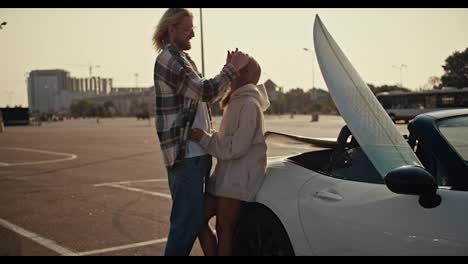 Close-up-shot-of-a-blond-guy-with-glasses-with-a-beard-in-a-plaid-shirt-hugs-his-blonde-girlfriend-and-strokes-her-hair,-a-girl-in-a-white-sweatshirt-looks-at-her-boyfriend-and-leans-on-a-white-convertible-in-which-there-is-a-white-surfboard-on-a-large-square-in-the-city