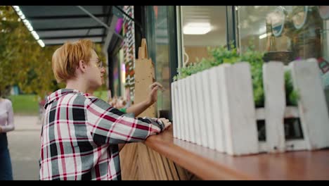 A-blonde-girl-with-a-short-haircut-in-a-checkered-shirt-and-glasses-and-her-girlfriend-with-a-short-green-haircut-in-a-Green-shirt-and-with-tattoos-take-hot-dogs-from-a-street-cafe-in-the-park-in-the-summer