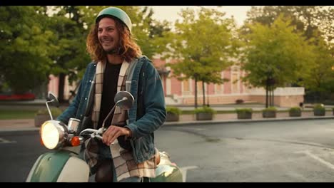 A-happy-brunette-guy-with-curly-hair-in-a-denim-jacket-drives-up-on-his-moped,-his-girlfriend-in-a-checkered-shirt-and-a-white-helmet-runs-up-to-him,-gets-on-the-moped-with-the-guy-and-they-leave