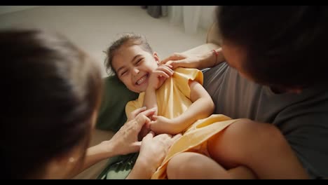 Close-up-shot-of-a-happy-little-girl-in-a-yellow-dress-laughing-and-rejoicing-while-being-tickled-by-her-parents-sitting-on-a-light-brown-in-a-modern-apartment