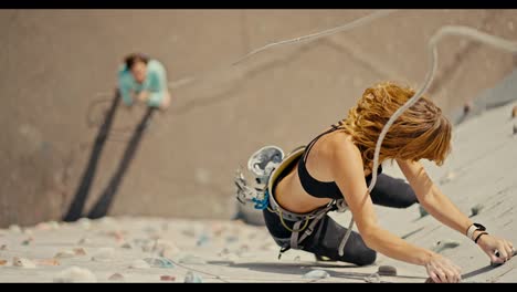 A-blonde-girl-in-a-black-sports-summer-uniform-climbs-up-a-white-climbing-wall-with-multi-colored-ledges-and-she-is-belayed-by-her-friend-in-a-blue-jacket-who-stands-on-the-ground-below-and-holds-a-rope.-Top-view-of-a-blonde-girl-climbing-up-a-climbing-wall-and-practicing-rock-climbing