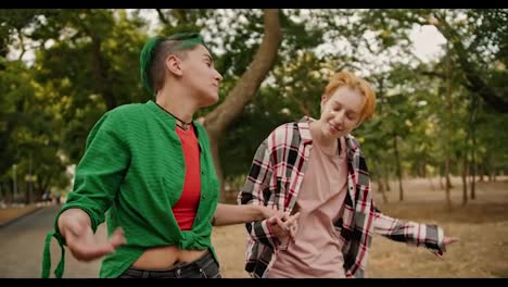 A-lesbian-girl-with-short-green-hair-and-a-green-shirt-and-her-blonde-girlfriend-with-short-hair-in-a-plaid-shirt-communicate-and-walk-hand-in-hand-and-shrug-their-shoulders-in-the-park-during-their-date-in-the-summer