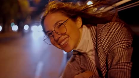 A-happy-and-cheerful-brunette-girl-in-round-glasses-smiles-broadly-in-a-gray-coat-and-looks-out-of-the-rear-window-of-a-modern-car-in-the-evening.-Happy-brunette-girl-looks-out-of-the-car-window-during-an-evening-drive-around-the-city