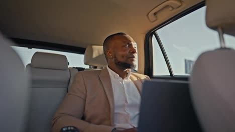 A-confident-brunette-man-with-Black-skin-and-a-beard-in-a-light-brown-shirt-holds-a-laptop-in-front-of-him-and-types-there-during-his-business-trip-in-a-modern-car-outside-an-urban-area